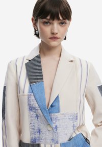 Patchwork blazer combining cream, blue, and striped panels. Features a deep V-neck, button closure, and contrast stitching. Textured fabric.