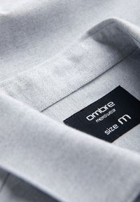 Gros plan sur le col d'une chemise gris clair avec une étiquette noire portant l'inscription « Ombre menswear taille M » cousue à l'intérieur de l'encolure.