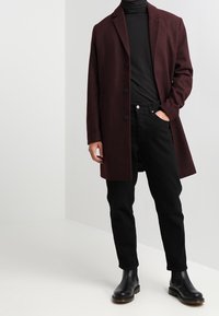 Cappotto bordeaux con revers a punte e bottoni neri, indossato sopra un dolcevita nero, abbinato a jeans neri e stivaletti neri.