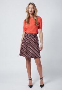 Oranges, geripptes Stricktop, kombiniert mit einem gemusterten schwarzen Rock, der geometrische Designs in Rot, Blau und Weiß aufweist. Das Modell trägt schwarze High Heels.