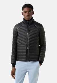 Sort dunjakke med chevron-vatteringsmønster, høj krave og lynlås foran. Har en lynlåslomme og ribstrikkede manchetter.