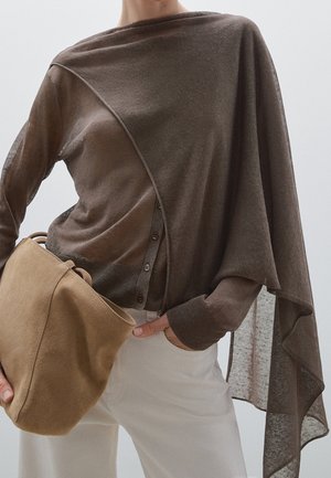 Person iført en brun let gennemsigtig asymmetrisk top og lyse beige bukser, som holder en stor ruskindstaske i tan-farve i hoftehøjde.