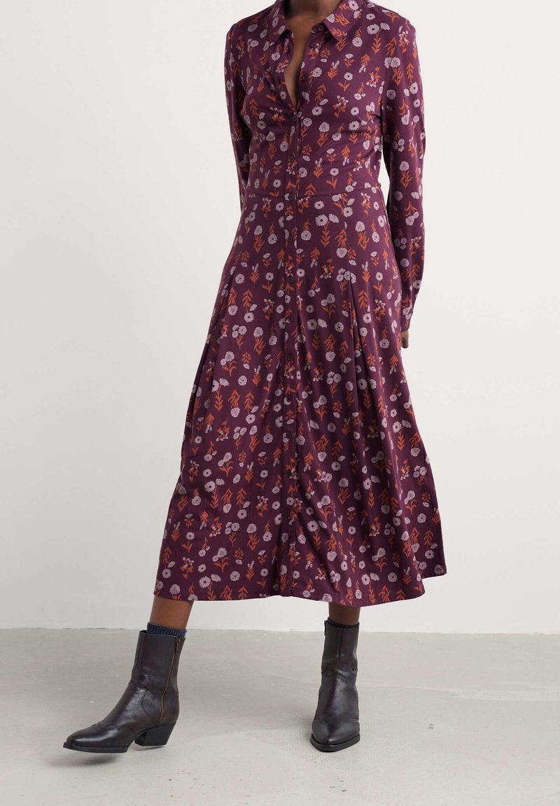 Robe longue à manches violettes avec imprimé floral orange et rose, boutonnée sur le devant, associée à des bottines noires et des chaussettes bleues.