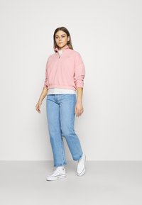 Sweatshirt rosa claro de pulôver com gola zip e barra canelada, combinado com jeans de perna reta azul claro e ténis brancos de cano alto.