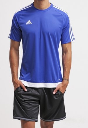 Mężczyzna w niebieskiej sportowej koszulce Adidas z białymi paskami na rękawach oraz czarnych sportowych spodenkach, z rękami w kieszeniach.