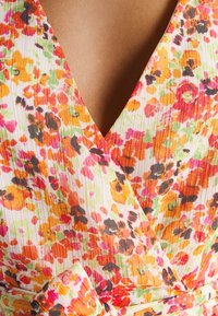 Robe portefeuille fleurie aux nuances d'orange, de rose et de vert sur un tissu blanc texturé, avec un décolleté en V et une taille ceinturée.