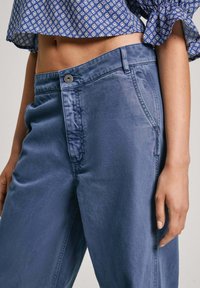 Lichtblauwe, katoenen broek in een ontspannen pasvorm. Voorzien van een knoopsluiting, zijzakken en subtiele branding op de tailleband.