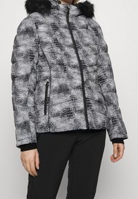 Wypełniona kurtka w biało-czarnym kolorze z wzorem imitującym skórę krokodyla, z zapięciem na zamek błyskawiczny oraz odpinanym kapturem z sztucznego futra. Gładka tekstura i dopasowany krój.