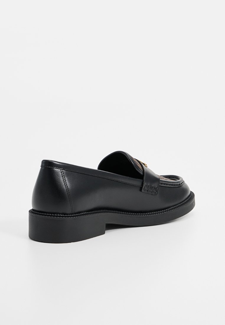 Zwarte leren instapper met dikke zool en decoratief metalen detail, van opzij weergegeven op een witte achtergrond.