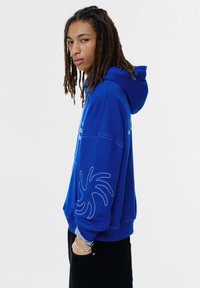 Blauwe hoodie met een spinnenwebontwerp op de mouw, gemaakt van zacht materiaal. Voorzien van een capuchon met trekkoord en geribbelde boorden. Zwarte broek.