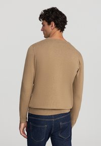 Maglione marrone a maglia con scollatura rotonda, polsini e orlo a costine, indossato sopra jeans di denim blu scuro, che mettono in mostra una texture morbida.