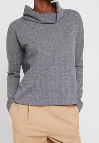 Grå stickad tröja med cowl-hals, långa ärmar och strukturerat mönster. Matchad med ljusbeige byxor som har en avslappnad passform.