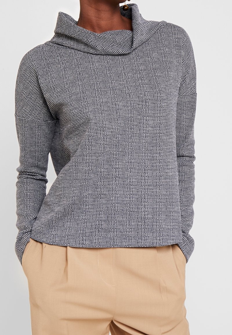 Grå stickad tröja med cowl-hals, långa ärmar och strukturerat mönster. Matchad med ljusbeige byxor som har en avslappnad passform.