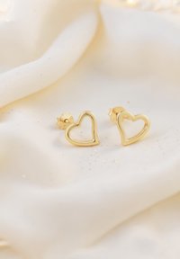 Orecchini a perno a forma di cuore in oro con centro vuoto, texture liscia e chiusura a farfalla, appoggiati su uno sfondo morbido e chiaro.