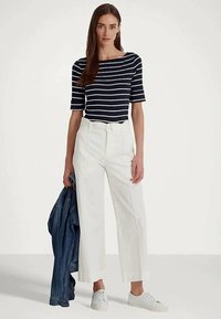 Γυναίκα φορώντας μια μαύρη-άσπρη ριγέ μπλούζα, ψηλόμεσες λευκές παντελόνες με ζώνη, λευκά αθλητικά παπούτσια, κρατώντας ένα τζιν μπουφάν με το ένα χέρι.