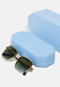 Melni saulesbrilles ar zeltītām akcentiem un tumši zaļām lēcām, kopā ar teksturētu gaiši zilu astoņstūra futrāli.