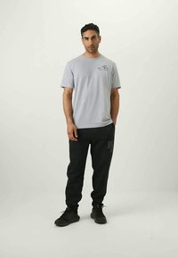 Man staat in een lichtgrijze T-shirt en zwarte joggingbroek met zwarte sneakers, handen ontspannen aan de zijkanten, neutrale achtergrond.
