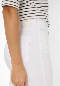 Jean en denim blanc avec une finition texturée, accents subtils sur les poches et étiquette crème. Présente un design ajusté et une taille mi-haute.