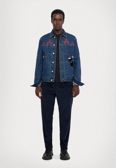 Denim jacket with red floral embroidery, classic collar, button closure, and two front pockets. Worn over a black shirt and navy pants.