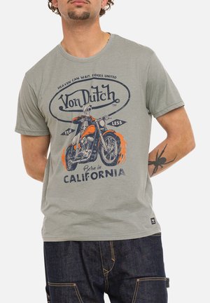 Homme portant un t-shirt gris Von Dutch avec un dessin de squelette conduisant une moto et le texte « Born in California » ainsi qu'un tatouage d'araignée sur le bras.