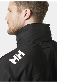 Czarna kurtka wierzchnia z wysokim kołnierzem, z wyraźnym białym logo na ramieniu i gładkim, teksturowanym materiałem.