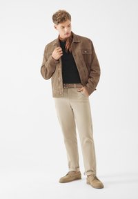 Hnědá semišová bunda s dvěma náprsními kapsami, černá košile, béžové kalhoty a světle hnědé boty; model pózuje se založenýma rukama do bundy.