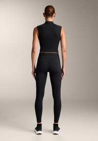 Črn športni crop top in prilegajoče hlače iz raztegljivega materiala, z visokim ovratnikom in brezšivno zasnovo. Črna obutev dopolnjuje videz.