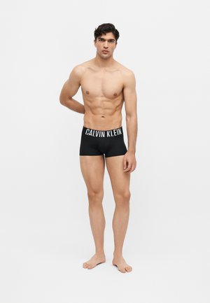 Mann med kort mørkt hår iført svarte Calvin Klein boxershorts, står barfot mot en ensfarget hvit bakgrunn.
