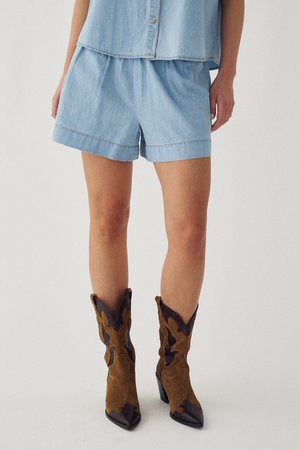 Persoon draagt lichtblauwe denim shorts en een bijpassend mouwloos shirt, gecombineerd met bruine en donkerbruine cowboylaarzen.
