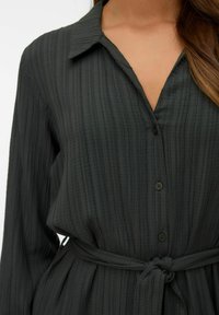 Chemise boutonnée vert foncé avec un tissu texturé et côtelé, un col et un détail de taille nouée. Caractéristiques de longues manches et de rayures verticales.