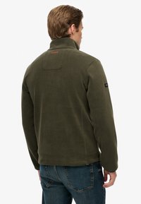 Zielona oliwkowa polarowa kurtka z wysokim kołnierzem, detalem na plecach i naszywką z logo na lewym ramieniu. Miękka faktura i dopasowany krój.