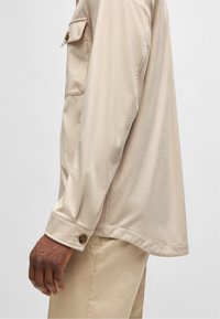 Beige overhemd met lange mouwen, een knoopmanchet en een patchzak. Glad materiaal en een ontspannen pasvorm, zijaanzicht tegen een neutrale achtergrond.