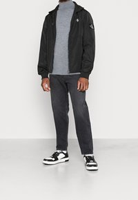 Veste noire à fermeture éclair avec capuche, pull à col roulé gris, jeans foncés et baskets noires et blanches. Logo notable sur la manche.