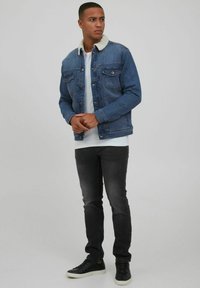 Chaqueta de denim con cuello de borreguito beige, en tela azul, con dos bolsillos en el pecho, llevada sobre una camiseta blanca, combinada con jeans negros y zapatillas negras.