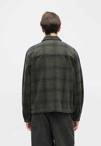 Temno zelena karirana srajca iz flanelne tkanine, z gumbi na sprednji strani, dolgimi rokavi in ravnim robom. Na hrbtni strani je prikazana zasnova yoke.