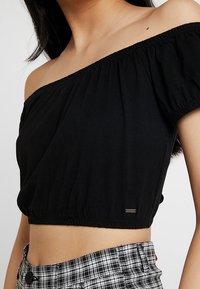 Svart avskuren topp utan axelremmar, gjord av mjukt material, med en elastisk halsringning och fåll. Anmärkningsvärd branding längst ner till vänster.