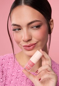 Botella de rubor en crema de color rosa con una tapa rosa claro, sostenida por una persona de piel suave, cabello oscuro y largo, que lleva un suéter rosa con textura.