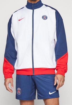 Personne portant une veste zippée Paris Saint-Germain blanche, bleu marine et rouge avec un short bleu marine assorti arborant les logos du club et de Nike.
