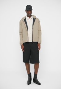 Beige gesteppte Jacke mit Reißverschluss, getragen über einem hellen Hemd und schwarzen Shorts. Schwarze Knöchelstiefel vervollständigen das Outfit.