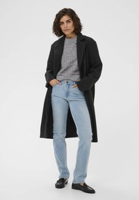 Manteau en laine noir sur un pull texturé gris, jeans bleu clair et mocassins noirs. Silhouette droite avec une coupe décontractée.