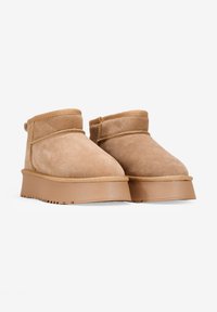 Botas de ante color marrón claro hasta el tobillo con puntera redondeada, suela de goma plana y detalles de costura mínimos. El diseño presenta un corte superior con puños.