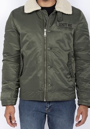 Homme portant une veste bomber verte avec col en shearling blanc, fermeture éclair devant, boutons, écusson avec le texte "Schott NYC," et jean bleu.