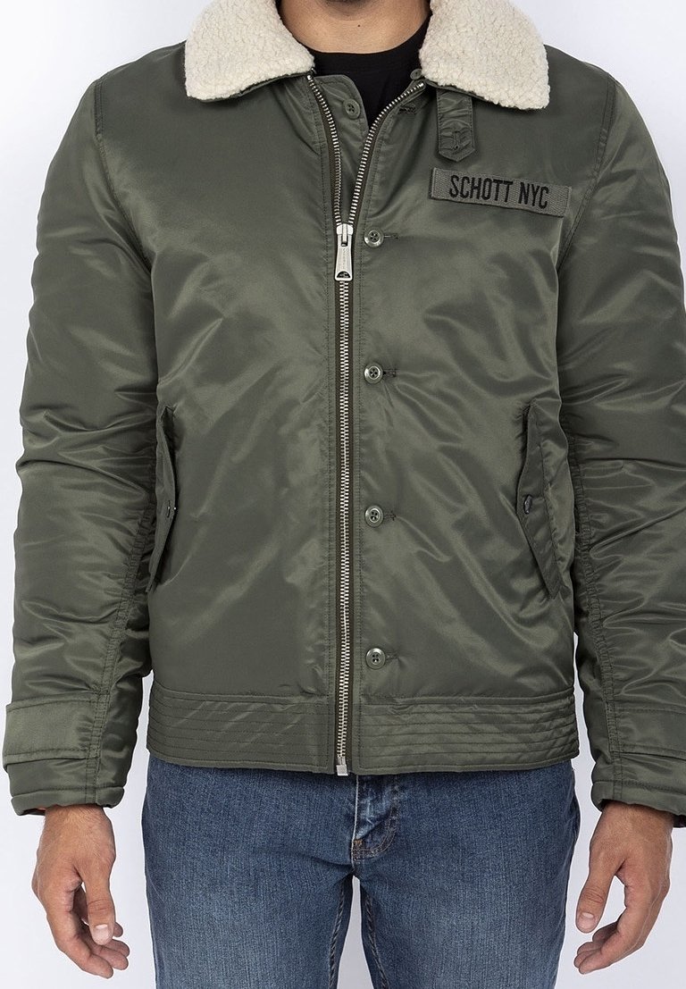 Homme portant une veste bomber verte avec col en shearling blanc, fermeture éclair devant, boutons, écusson avec le texte "Schott NYC," et jean bleu.