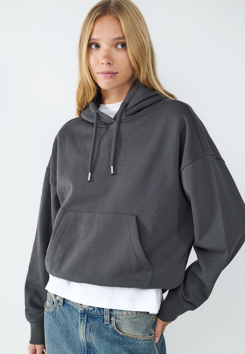 Junge Frau trägt einen dunkelgrauen, kurz geschnittenen Hoodie mit Fronttasche und Kordeln, darunter ein weißes Hemd und blaue Jeans.