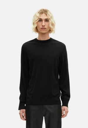 Blonde persoon met golvend haar draagt een eenvoudig zwart langärmige trui en zwarte broek, staand tegen een witte achtergrond.