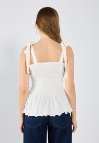 Bílý top s peplum střihem, s nařaseným trupem, nastavitelnými ramínky a volánkovým lemem. Měkká látka a volný design doplňují džínové spodní díly.