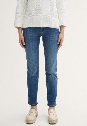 Slim-Fit blaue Jeans getragen mit einem cremeweißen strukturierten Strickpullover und cremefarbenen klobigen Loafers vor hellem Hintergrund.