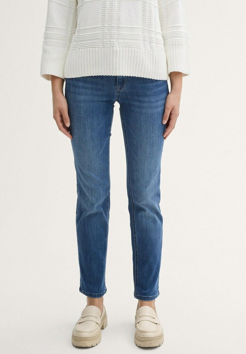 Jean slim bleu porté avec un pull en maille texturée écru et des mocassins épais couleur crème sur un fond clair.