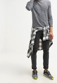 Grå texturerad långärmad skjorta, svart rutigt flanell skjort anknuten i midjan, smala svarta byxor och grå sneakers med gula accenter.