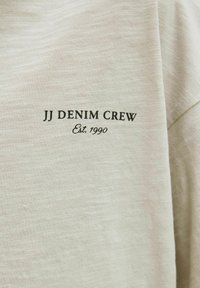 Lichtbeige t-shirt met "JJ DENIM CREW" in zwarte tekst en "Est. 1990" eronder. Zachte, gestructureerde stof met een ontspannen pasvorm.